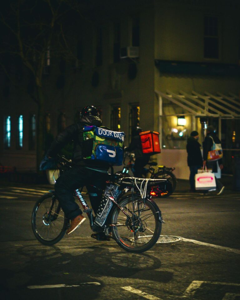 A dynamic, motion-blurred image of a delivery rider wearing a white helmet, an orange jacket, and dark pants, speeding on a silver scooter-motorcycle through illuminated city streets at night.