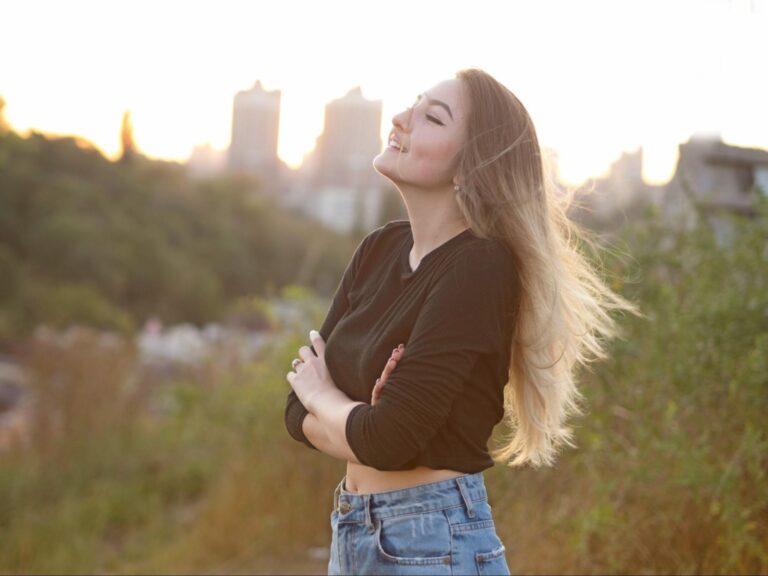 Confident young woman standing in natural sunlight with eyes closed and a peaceful smile, arms gently wrapped around herself—symbolizing self-acceptance, emotional healing, and inner worth.