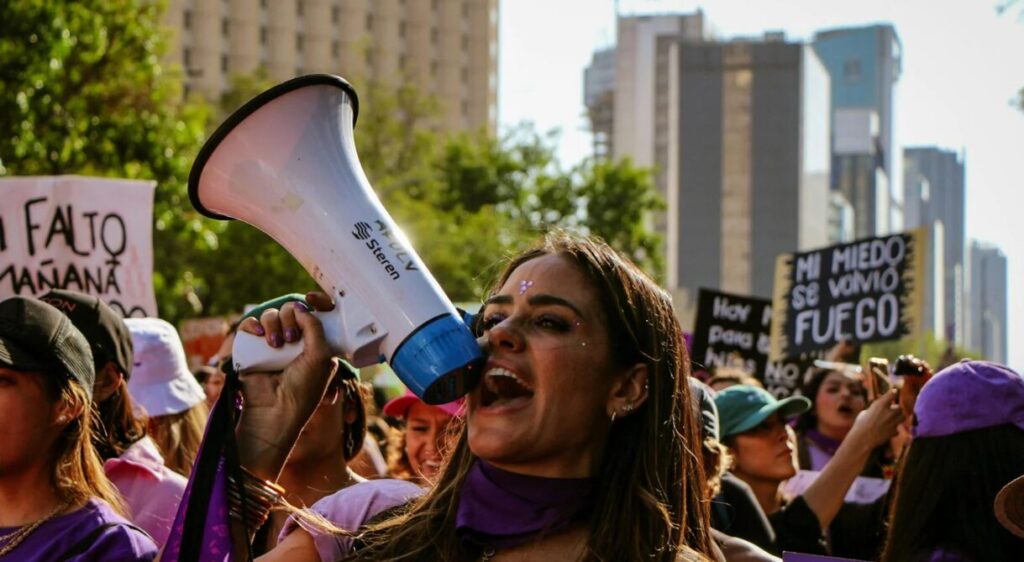A woman passionately shouting into a megaphone during a feminist protest march, surrounded by a crowd of women holding signs with empowering messages in Spanish. One visible sign reads "Mi miedo se volvió fuego" ("My fear turned into fire"). The atmosphere is energetic and defiant, with participants wearing purple and standing against a backdrop of urban high-rise buildings.)