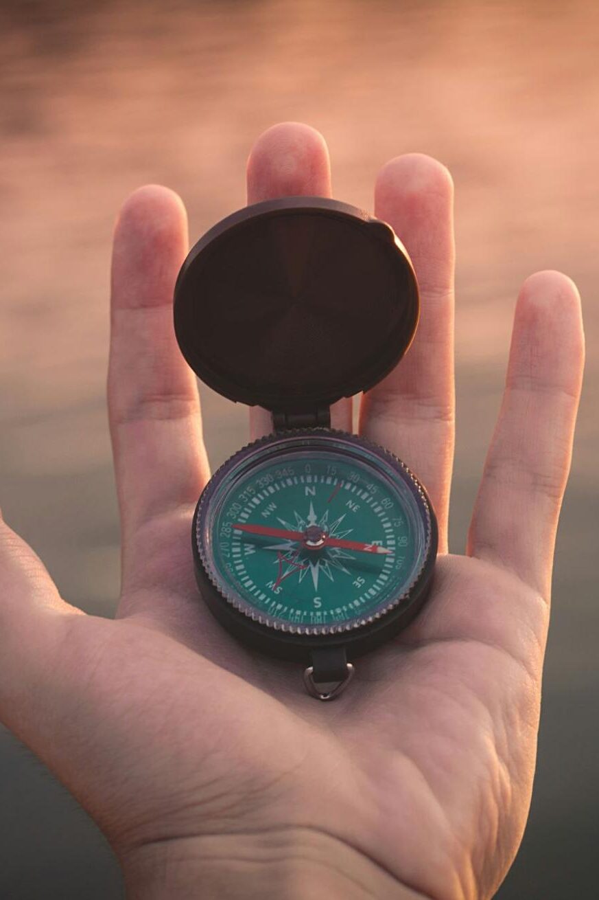 A hand holds an open, green compass with a visible needle, against a blurred background of water and warm light, possibly a sunset or sunrise over a lake.