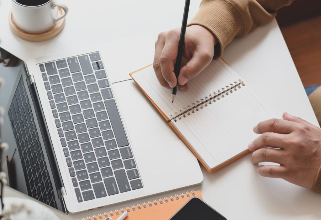Person writing notes beside laptop, organizing tasks to manage time well.