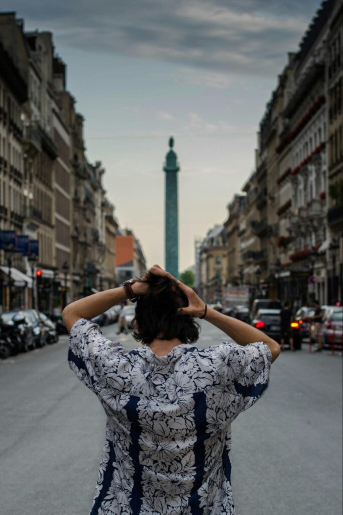 A person with dark hair, seen from behind, stands in the middle of a city street, looking towards a tall, slender monument in the distance. Buildings line both sides of the street, and cars are visible.