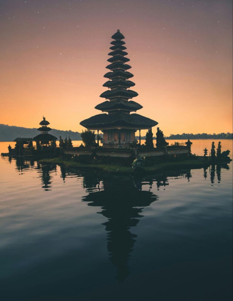 A serene sunrise or sunset scene features a multi-tiered Hindu temple reflected perfectly in the still water of a lake, with a soft orange and pink sky in the background.