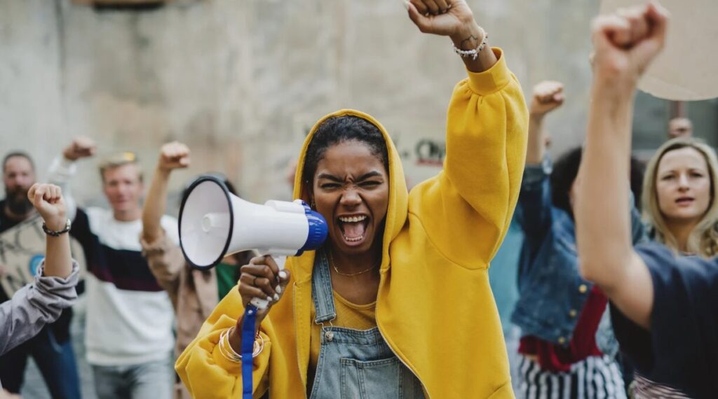 A young woman in a yellow hoodie passionately shouting into a megaphone during a protest, raising her fist in the air. She is surrounded by a diverse group of demonstrators, all with raised fists, expressing solidarity and determination. The mood is intense and empowering, capturing the spirit of activism and collective resistance.