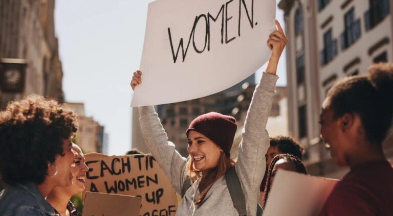 A young woman wearing a maroon beanie and grey hoodie joyfully holds up a large sign that reads “WOMEN” during a street protest. She is surrounded by a diverse group of women, smiling and engaging in the rally. Other protest signs are visible in the background, set against a backdrop of urban buildings, reflecting a powerful moment of feminist solidarity and celebration.