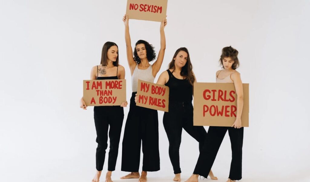 Four women standing barefoot against a plain white background, each holding handmade cardboard signs with bold red feminist messages. The signs read: "I AM MORE THAN A BODY," "NO SEXISM," "MY BODY MY RULES," and "GIRLS POWER." They wear neutral-toned tops and black pants, embodying strength, unity, and defiance in a minimalist yet powerful protest portrait.