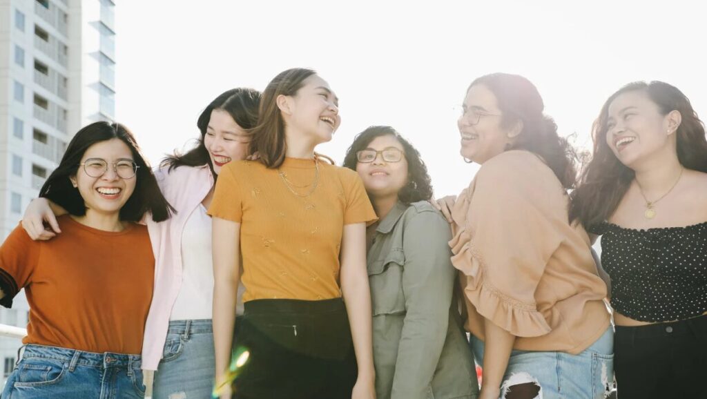 joyful group of six young women standing outdoors in sunlight, laughing and embracing each other.