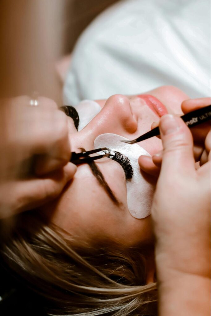 A professional uses two pairs of tweezers to apply eyelash extensions to a client lying down with white pads under her eyes.