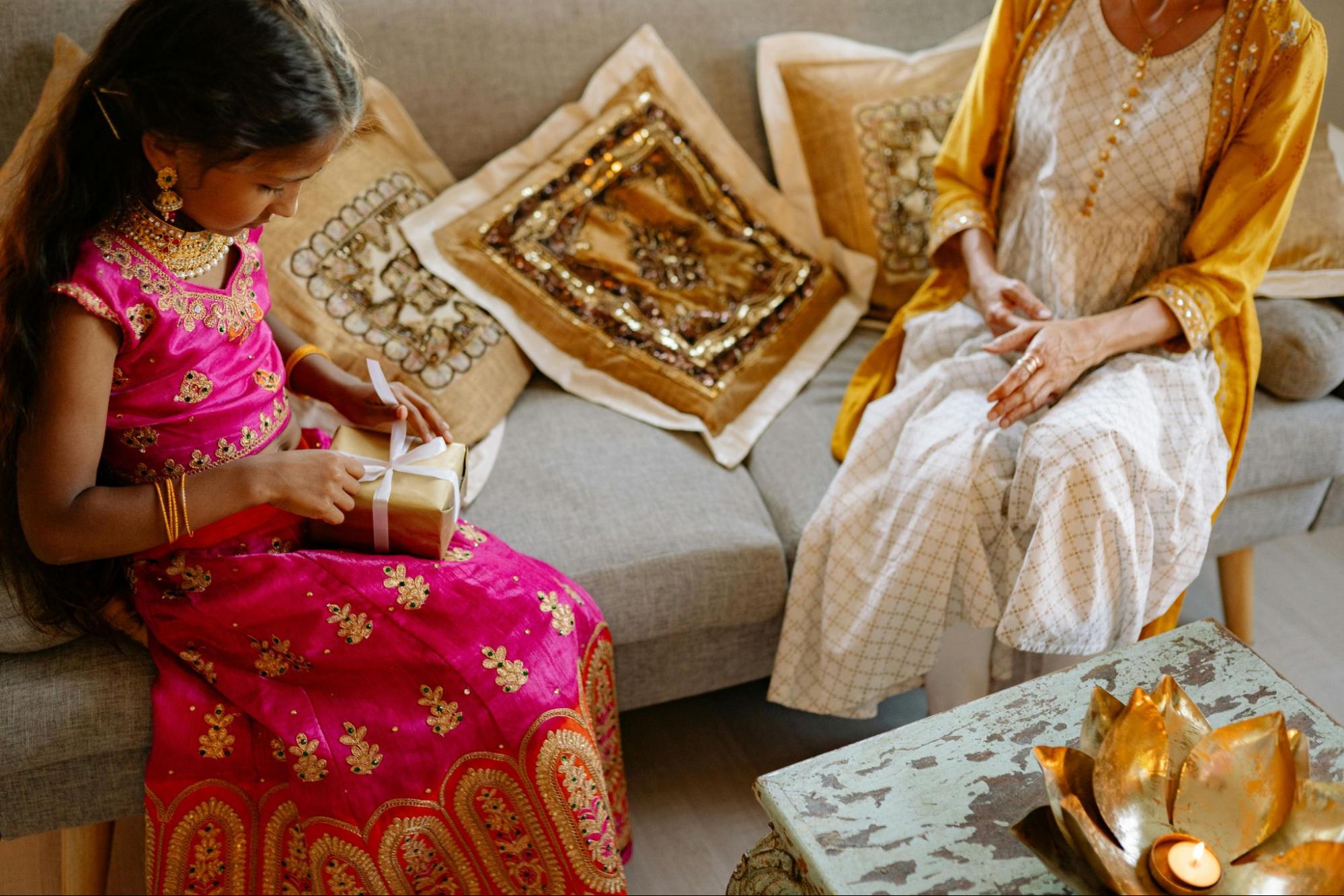 A young girl in a bright pink and gold traditional Indian dress is holding a wrapped gift with a white ribbon while sitting on a couch.