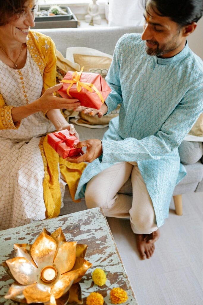 A woman in a yellow traditional Indian dress is handing a gift with a red bow to a man in a light blue traditional Indian shirt.