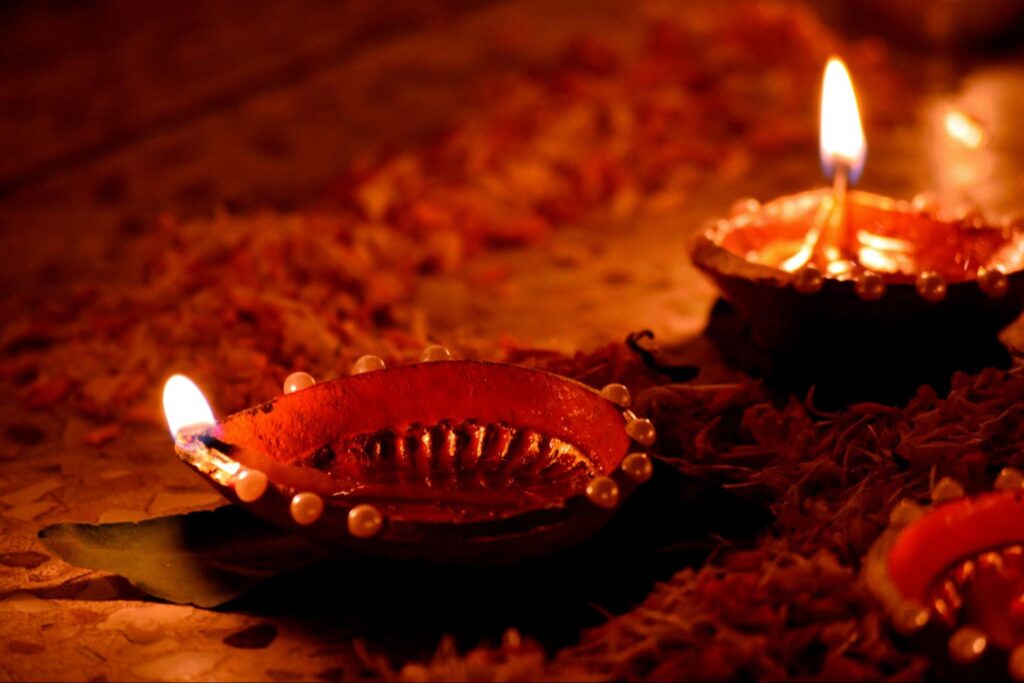 Two decorated clay lamps (diyas) with glowing wicks sit on a surface scattered with flower petals, with a third diya partially visible in the corner.