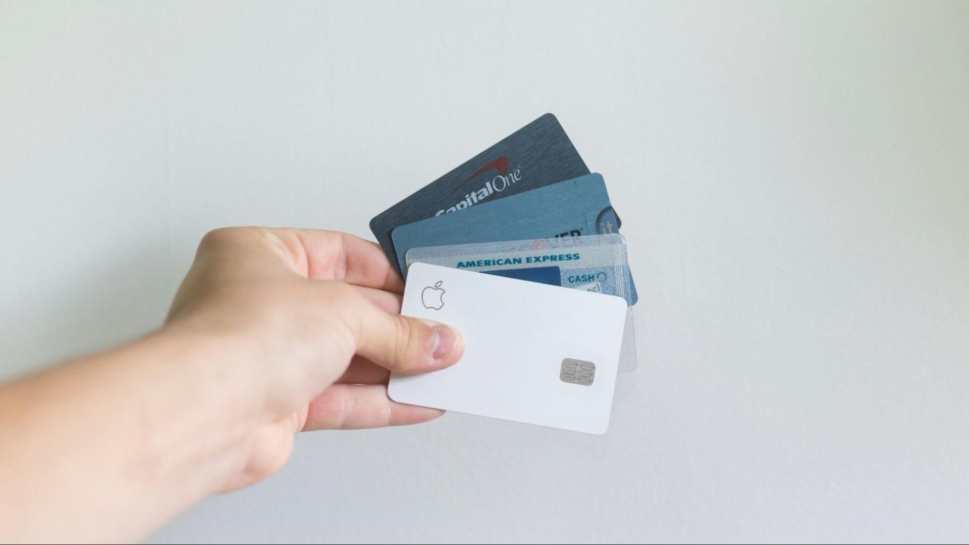 A hand holding a fanned-out stack of credit cards against a plain white wall, with a white Apple Card in the front.